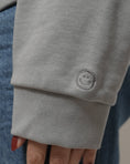 Load image into Gallery viewer, Close-up of a gray sweatshirt with a smiley face emblem on the sleeve. - Thread Vision Spotlight, Loe Hayley