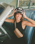 Load image into Gallery viewer, Woman sitting in a vintage car, wearing a black cap and brown fanny pack and smiling. - Thread Vision Client Spotlight, RESET by Serena Pitt