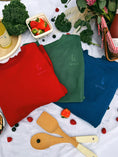 Load image into Gallery viewer, Three colorful aprons (red, green, blue) with a brand logo on a tablecloth with food and utensils. - Thread Vision Spotlight, Ignited Apparel
