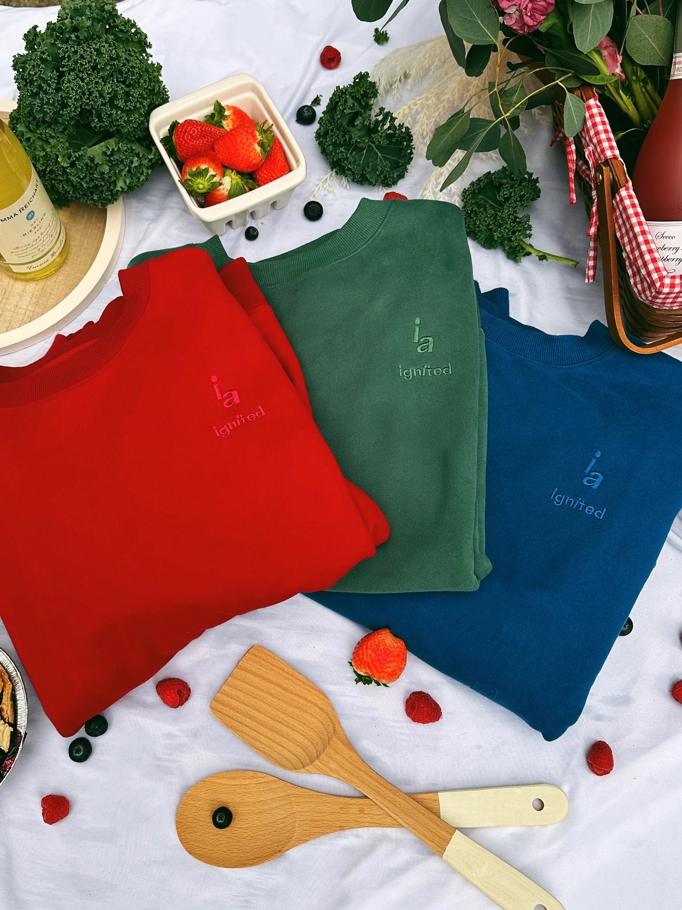 Three colorful aprons (red, green, blue) with a brand logo on a tablecloth with food and utensils. - Thread Vision Spotlight, Ignited Apparel