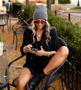 Load image into Gallery viewer, Woman sitting outdoors using a phone, wearing a black coat and gray beanie. - Thread Vision Spotlight, Hill x Sarah, Sarah Hill 
