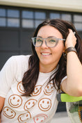 Load image into Gallery viewer, Woman with glasses and a smiley face shirt leaning against a wall. - Thread Vision Spotlight, Remington Avenue
