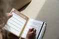 Load image into Gallery viewer, Person writing in a spiral-bound planner with January 2025 on a beige surface - Thread Vision Spotlight, Move Like Morgan
