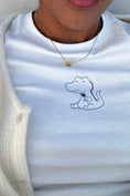 Load image into Gallery viewer, White t-shirt with a small dinosaur design on a person wearing a gold necklace. - Thread Vision Spotlight, Ignited Apparel

