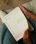 Load image into Gallery viewer, Person holding a paper planner notepad with a schedule on a couch - Thread Vision Spotlight, Ignited Apparel
