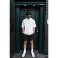 Load image into Gallery viewer, Man standing in front of a dark green door wearing a white t-shirt, black shorts, and white sneakers. - Thread Vision Spotlight, Kinda Handsome
