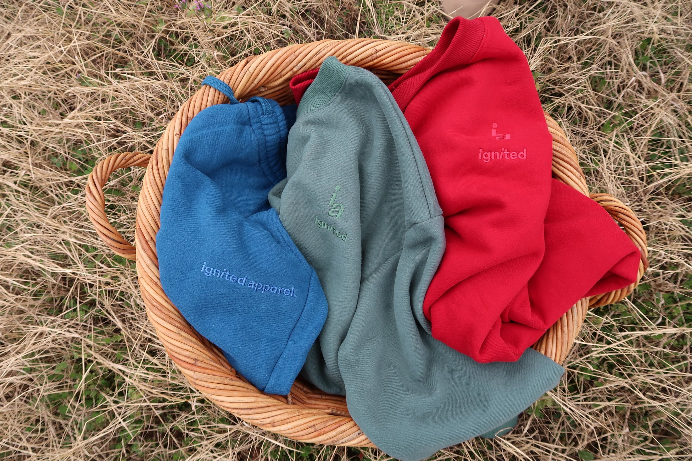 Three hoodies in blue, green, and red on a woven basket with 'ignited apparel' text against a natural background. - Thread Vision Spotlight, Ignited Apparel