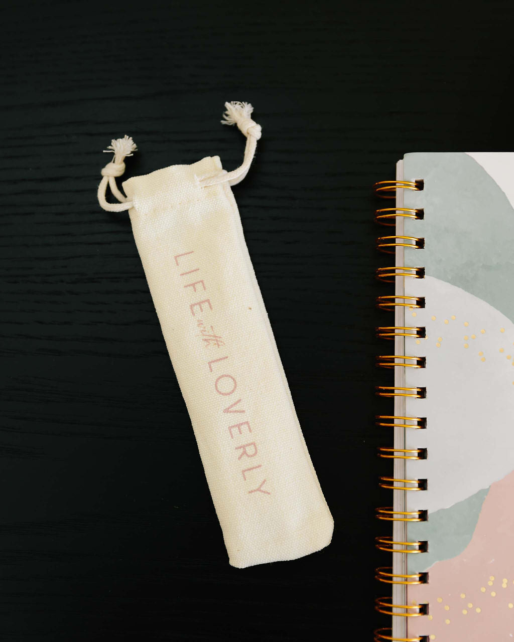 White drawstring pouch with 'LIFE is LOVELY' text next to a spiral-bound notebook on a dark surface. - Thread Vision Spotlight, Life With Loverly