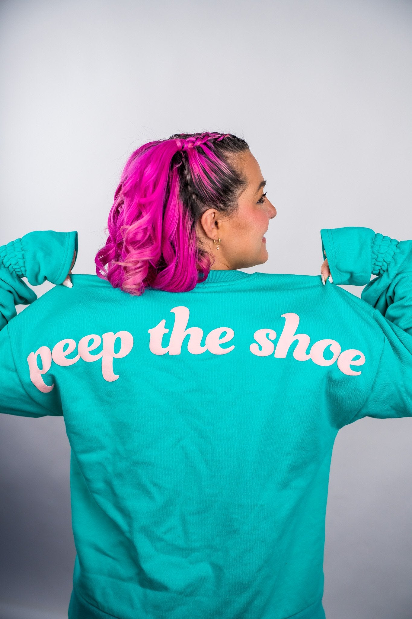 Person wearing a teal jacket with 'peep the shoe' text on the back against a plain background - Thread Vision Client Spotlight, 10/10 Deanna Giulietti 