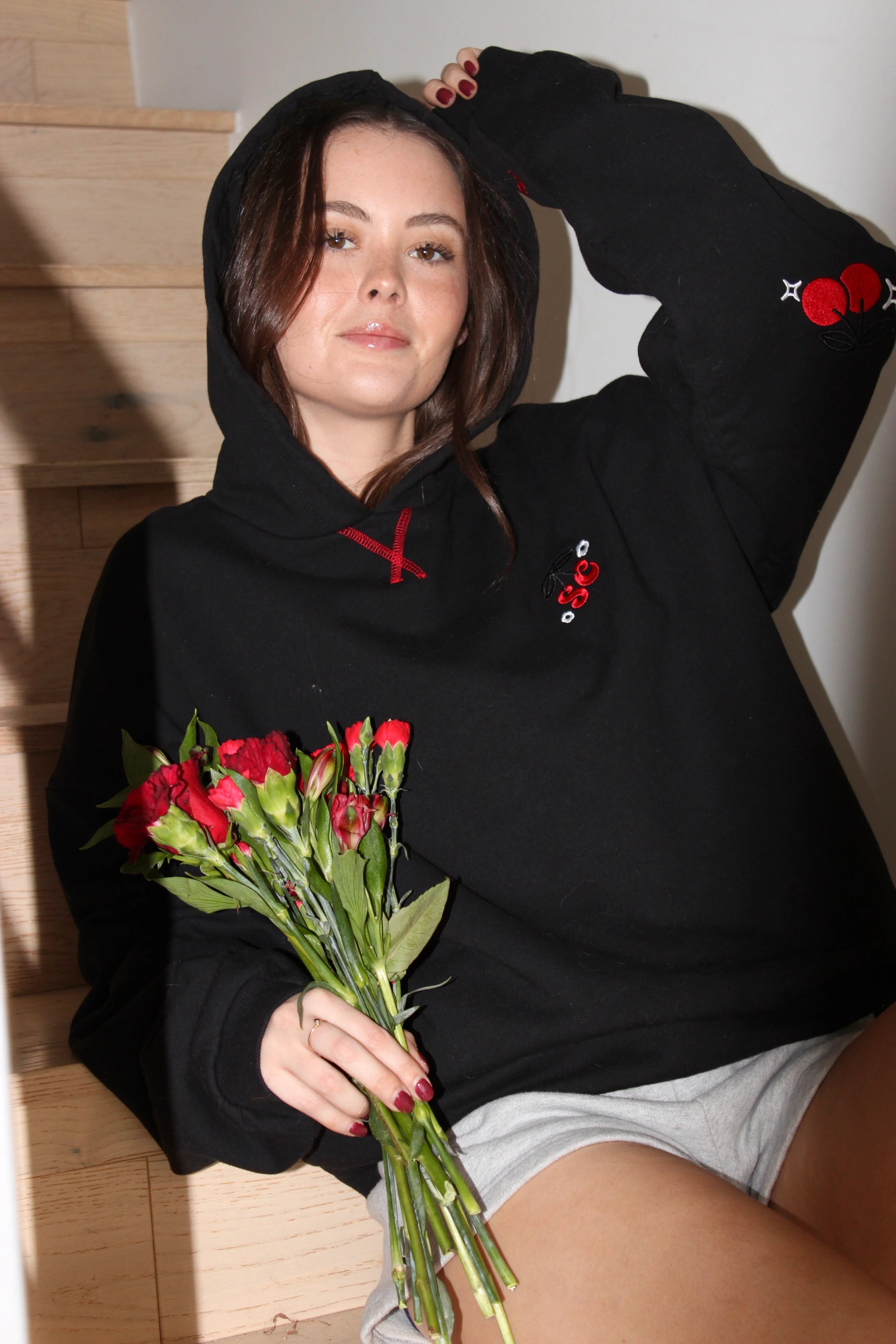 Person wearing a black hoodie with red floral designs, holding flowers indoors. - Thread Vision Client Spotlight, Sunday Cherries