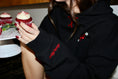 Load image into Gallery viewer, Person holding a red cupcake with white frosting, wearing a black hoodie with red text. - Thread Vision Client Spotlight, Sunday Cherries
