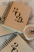 Load image into Gallery viewer, Two spiral-bound notebooks with '2024' design and a coffee cup on a marble surface. - Thread Vision Client Spotlight, Skinny Hangover
