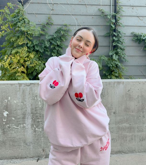 Person wearing a pink outfit with floral designs against a gray wall with greenery. - Thread Vision, Client Sunday Cherries