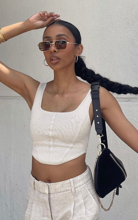 Woman wearing a white sleeveless top and light-colored pants, holding a black handbag against a plain background. - Thread Vision, Client Asha Bailey