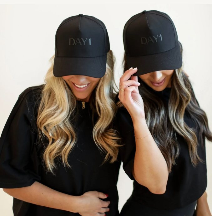 Two women wearing black caps with 'DAY1' text on a plain background -   Thread Vision, Client Day 1 Fitness