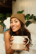 Load image into Gallery viewer, Woman wearing a brown beanie and holding a mug in a cozy indoor setting with plants. - Thread Vision Spotlight, Ignited Apparel
