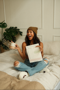 Load image into Gallery viewer, Woman sitting on a bed holding a coffee cup and a notepad with a plant in the background. - Thread Vision Spotlight, Ignited Apparel
