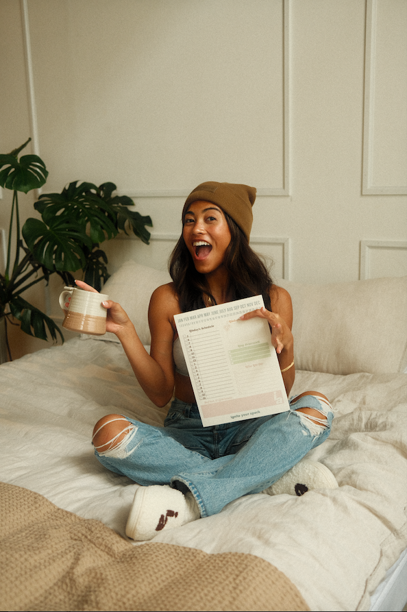 Woman sitting on a bed holding a coffee cup and a notepad with a plant in the background. - Thread Vision Spotlight, Ignited Apparel