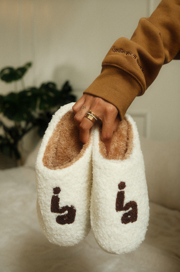 White slippers with brown interior and 'ia' branding held by a person wearing a brown sweatshirt. - Thread Vision Spotlight, Ignited Apparel