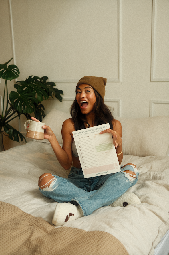 Woman sitting on a bed holding a coffee cup and a notepad, with plants in the background - Thread Vision Spotlight, Ignited Apparel