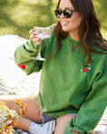 Load image into Gallery viewer, Woman wearing a green sweatshirt with cherry designs, holding a glass outdoors. - - Thread Vision Client Spotlight, Sunday Cherries
