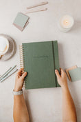 Load image into Gallery viewer, Green notebook held by hands on a light surface with stationery items. - Thread Vision Spotlight, PCOS Planner
