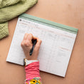 Load image into Gallery viewer, Person writing in a meal planner on a wooden surface with a green towel. - Thread Vision Spotlight, Oh Snap Macros
