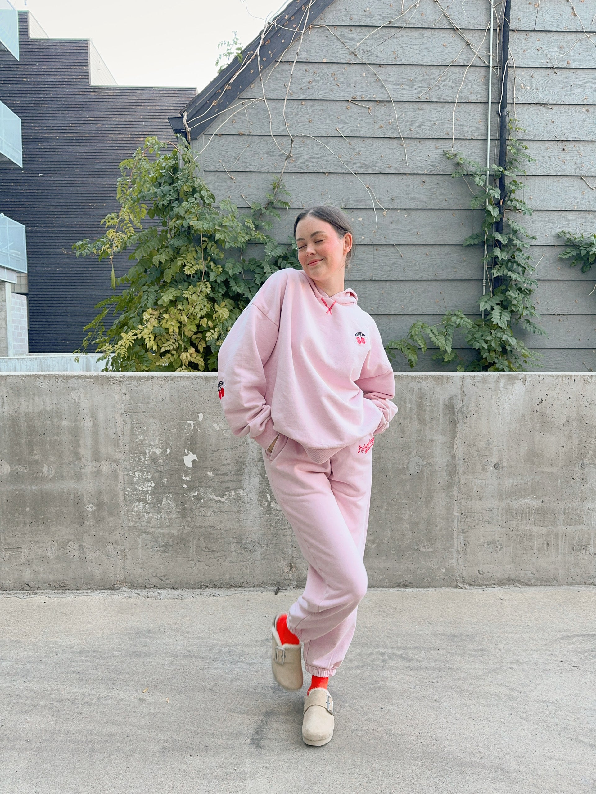 Person wearing a pink tracksuit standing outdoors on a concrete surface with a gray wall and plants in the background. - - Thread Vision Client Spotlight, Sunday Cherries